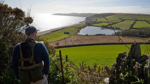 This landscape photograph captures Beesands on the coast of Devon, England, United Kingdom, during a late winter morning. In the foreground, a person identified as Jamie Weinbren is looking out over the scenery, equipped with tactical outdoor gear. The image features lush green fields, a patchwork of farmland, and the freshwater lake known as Widdicombe Ley. Beyond the fields, the coastline stretches towards the village of Beesands and the vast expanse of the sea. The distant headland of Start Point is visible on the horizon, a notable landmark along the coastline. The light from the morning sun creates a reflective sheen across the water, illuminating the rolling countryside and coastal features typical of this scenic part of Devon.